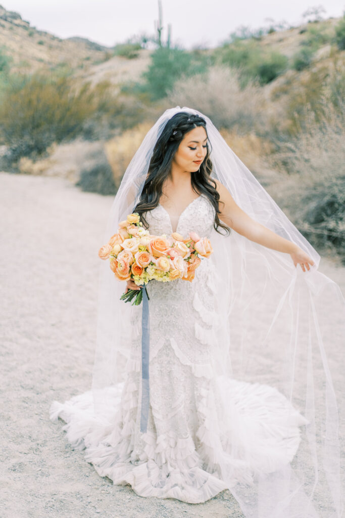 Bride walking in desert landscape holding out veil to side and looking down and holding bouquet.