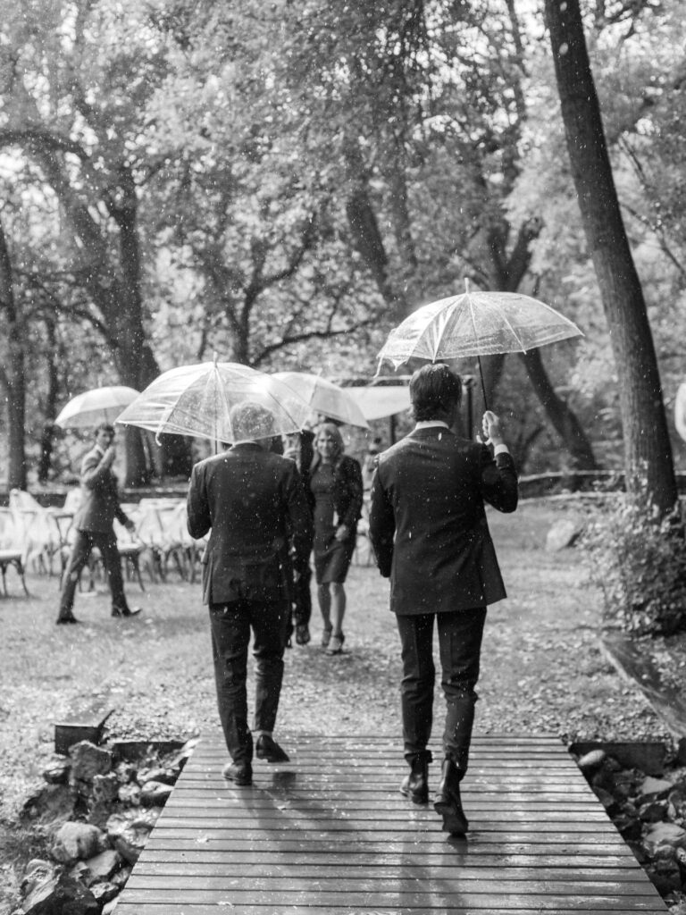 Men waling over wooden bridge holding umbrellas in the rain to wedding ceremony.