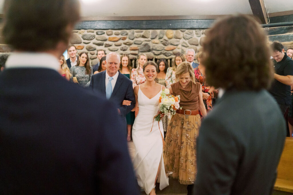 Bride walking between mother and father in aisle to groom at wedding ceremony.
