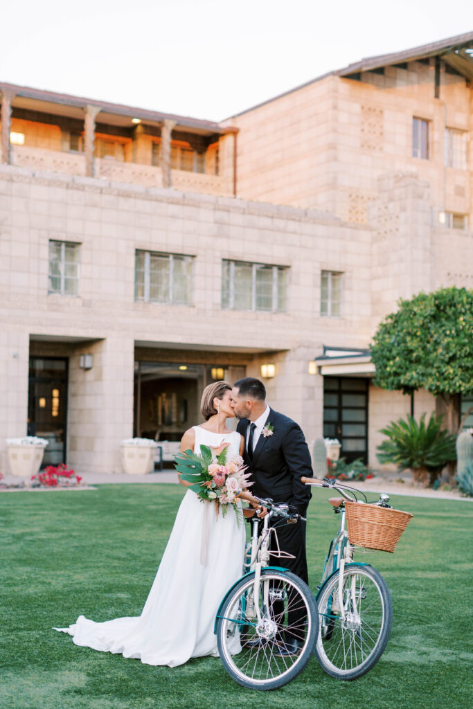Bride and groom kissing on grass in front of Arizona Biltmore with bikes and a bouquet in front of them.