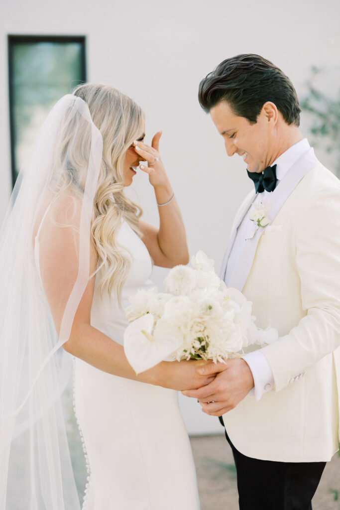 A man and a woman, the woman holding a bouquet and the man's hand on hers. The bride is wiping her eye from tears.