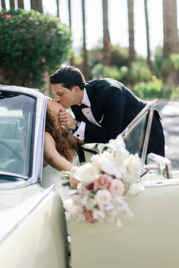 Groom kissing bride who is sitting in driver seat of vintage car.