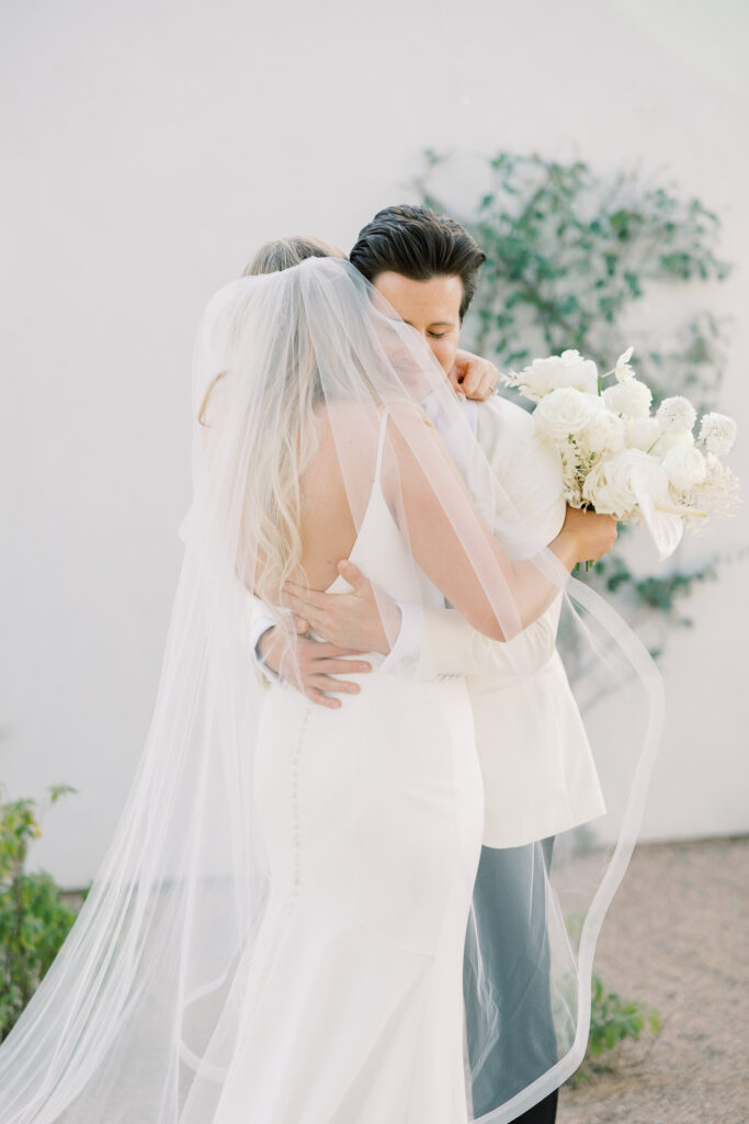 A man in tux and woman in wedding dress hugging in front of white wall.