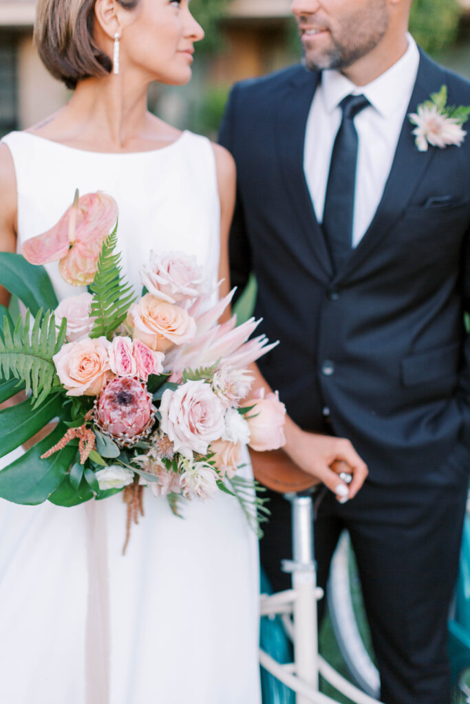 Tropical styled wedding bride and groom with bouquet, standing with a bike.
