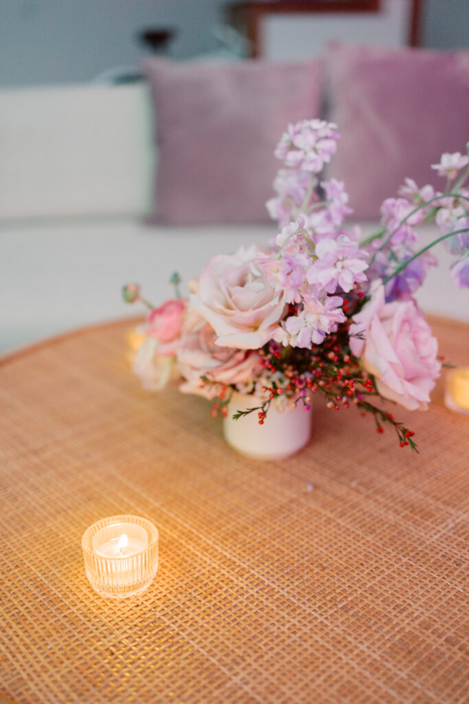 Table with centerpiece of pink, white, red, and purple flowers and a votive candle with a white couch in the background with pink pillows in the background.