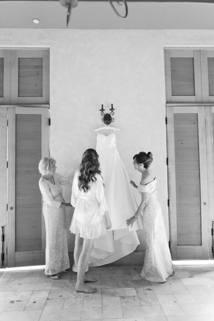 Bride looking at wedding gown hanging on wall with two other women.