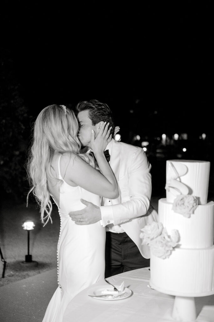 A black and white photo of a man and woman kissing in front of a cake that's on a table at night.