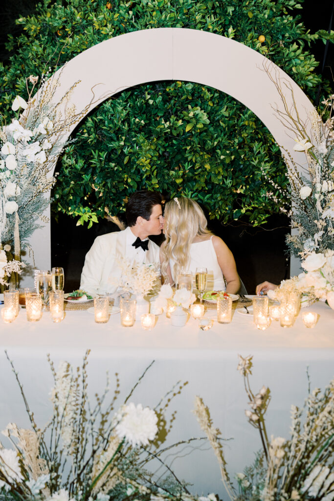 A man and woman kissing in front of an arch with white flowers on it with an orange tree behind the arch. Flowers in front of the table on ground.