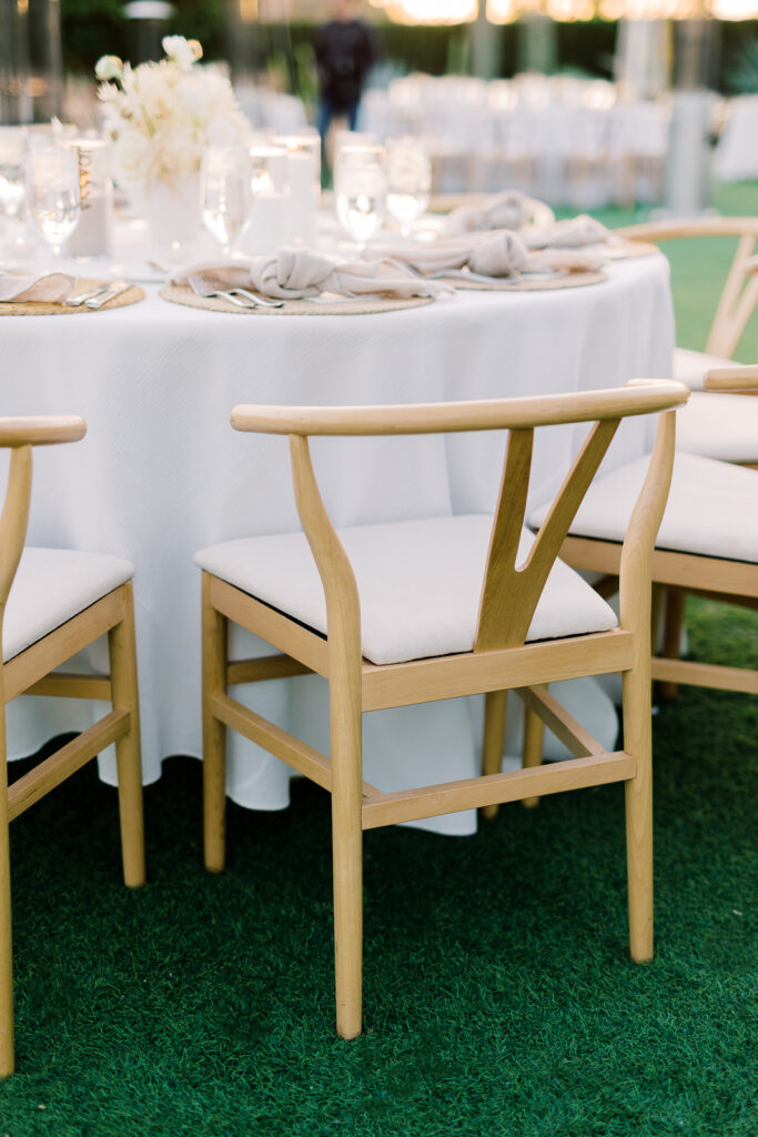 A tan chair with a white cushion at a round table set with white table cloth, place settings, candles and a centerpiece.
