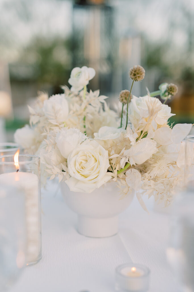 Centerpiece with roses cups and candles and white table cloth underneath it.