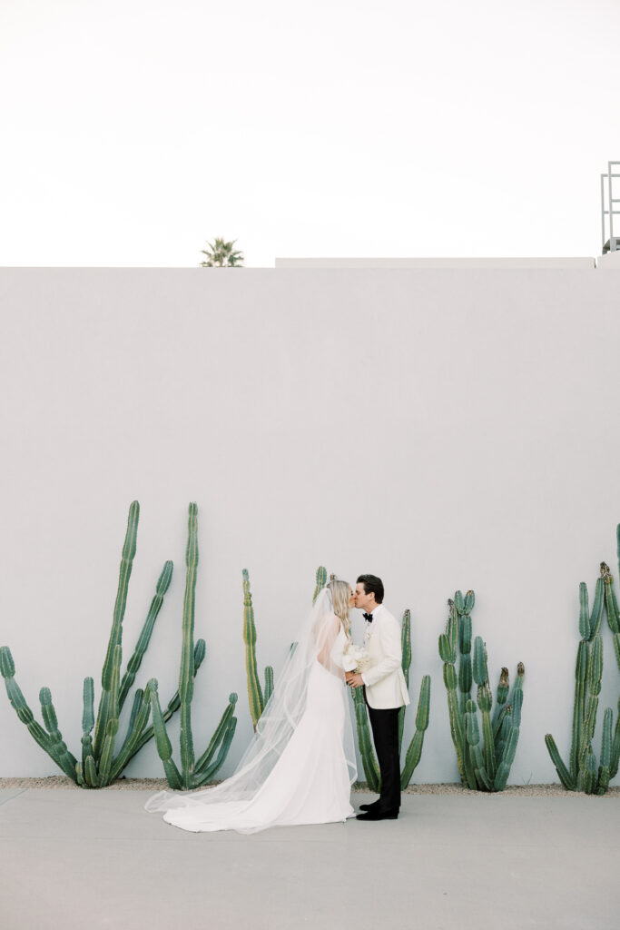 A man and woman kissing kissing with cacti behind them and a white wall behind the cacti.