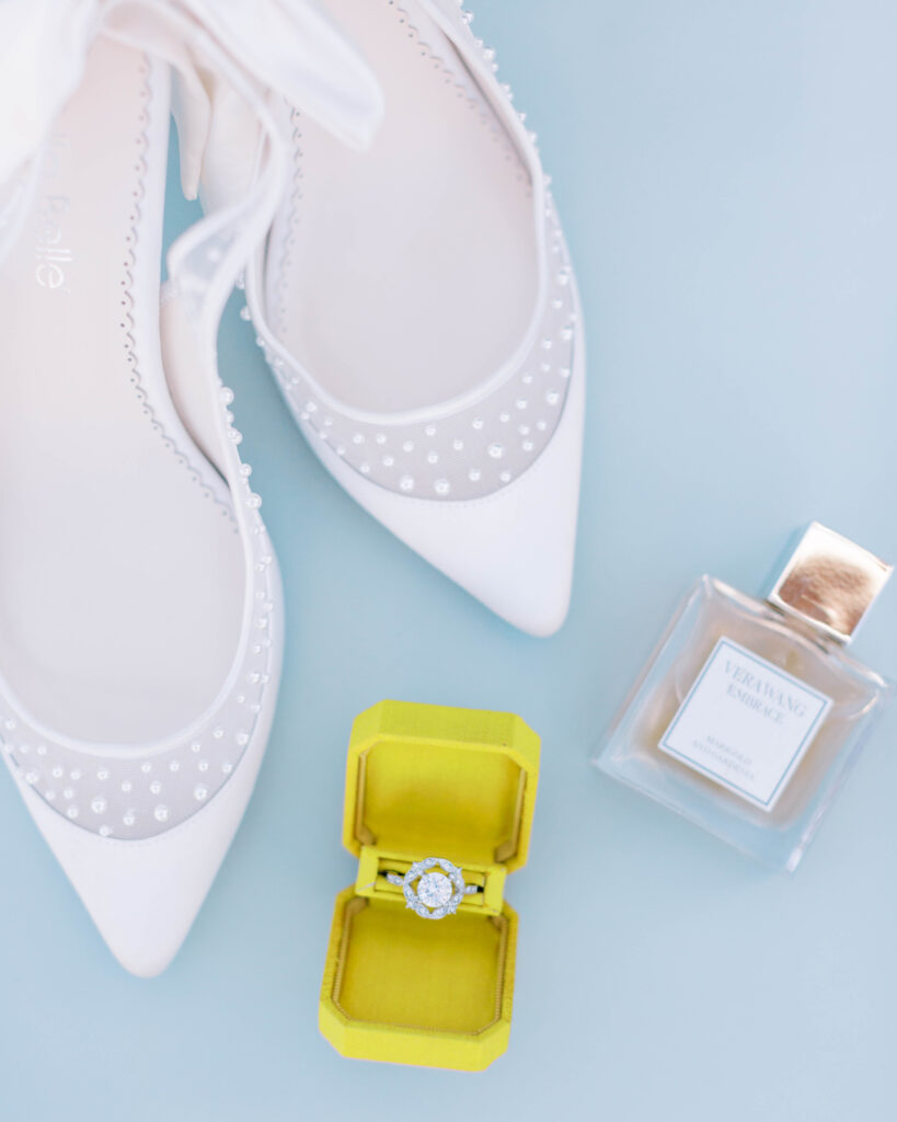 Bride details of perfume, ring in yellow box, and white wedding shoes.