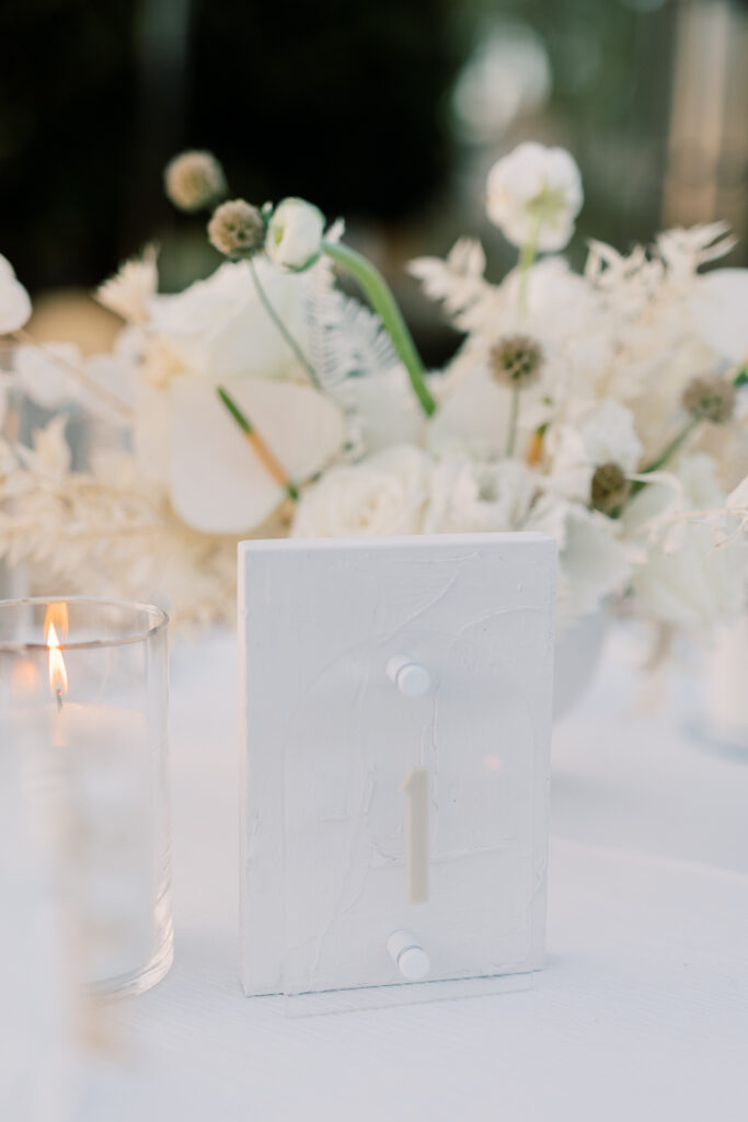 A white table number with flowers behind it and candle beside it and white table cloth underneath.