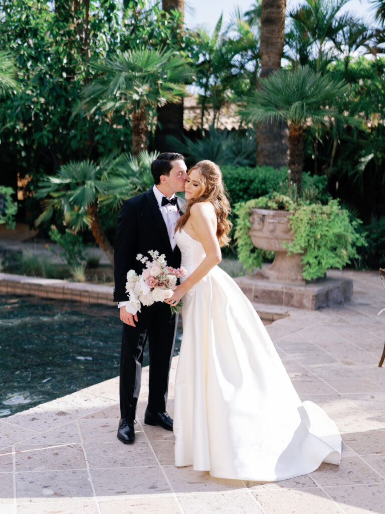 Groom kissing bride on the cheek in front of pool and greenery at Royal Palms.