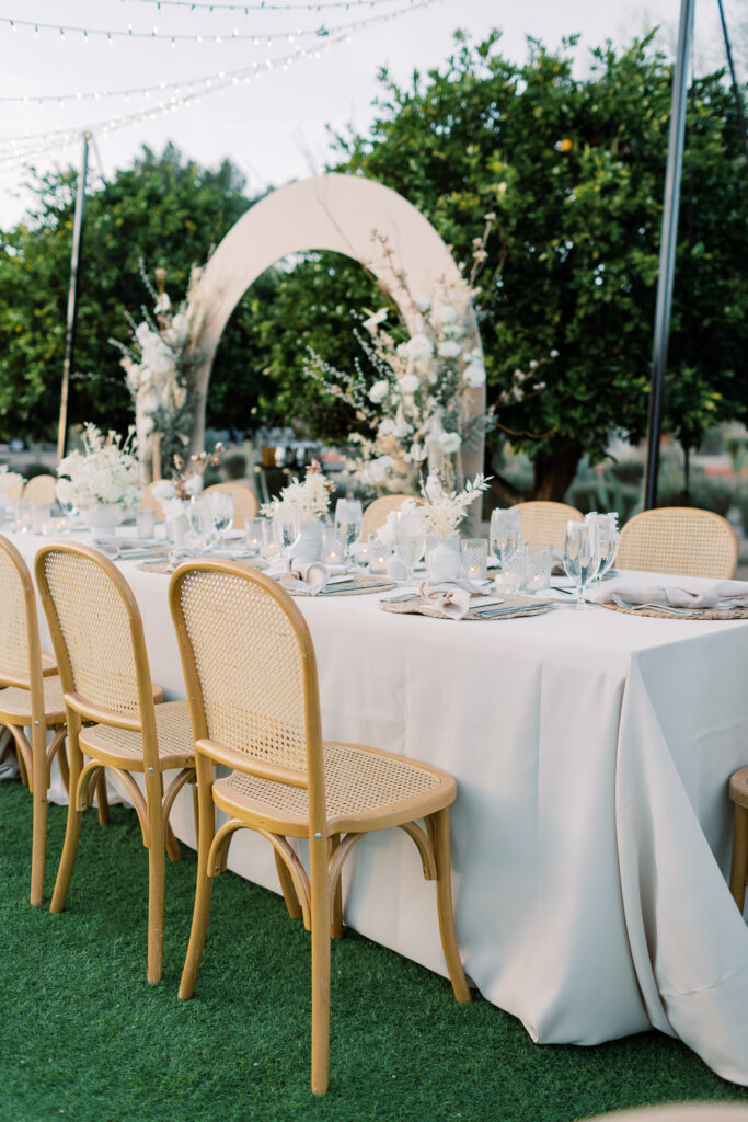 Long reception table with an arch and flowers placed behind it and orange trees in the backgound.