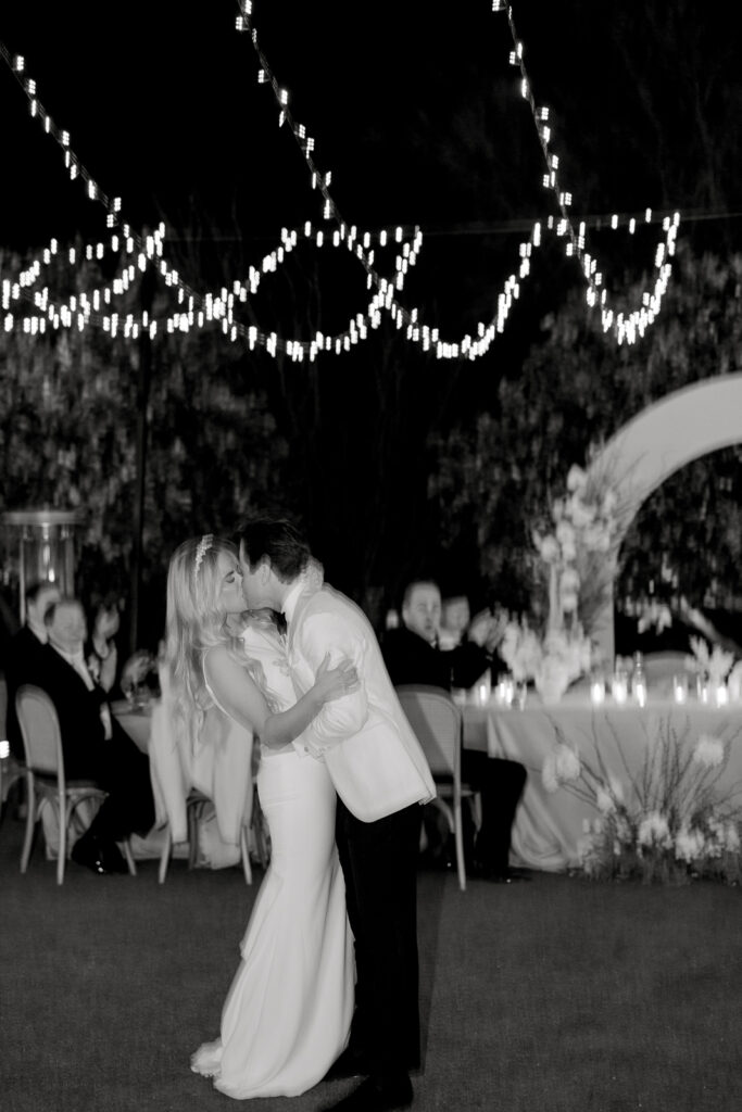 A man and woman kissing in the dark with lights attached to a wire and an arch in the background with trees behind it.