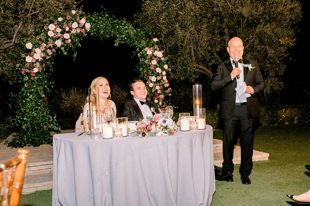 Bride and groom smiling while seated at sweetheart table at evening wedding reception. Man standing next to table holding microphone.