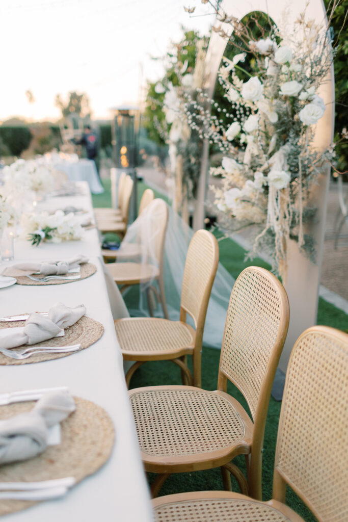 A bunch of white flowers, a line of brown chairs and utensils and napkins on a long table with a clear sky overhead.
