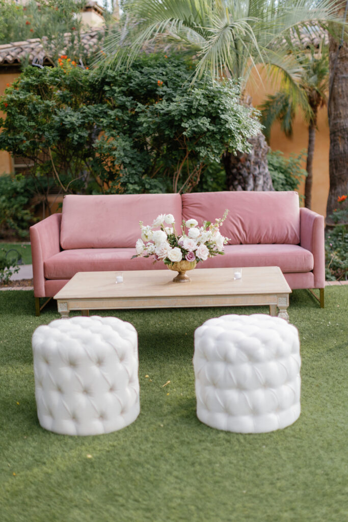 Wedding lounge area with pink couch, flower centerpiece on coffee table and two white apolstered stools.