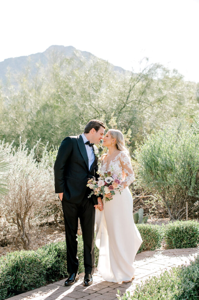 Bride and groom standing on brick path in desert landscape touching noses.