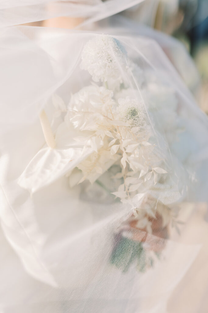White flowers wedding bouquet with bride in white dress behind it and veil covering flowers.
