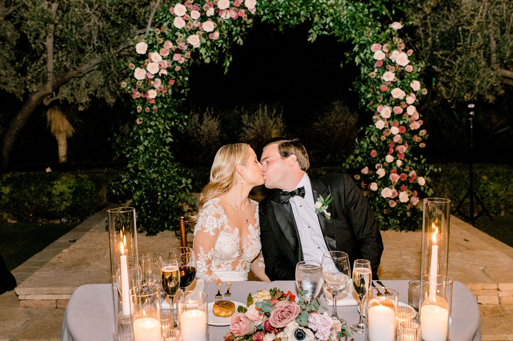 Bride and groom kissing while seated at sweetheart table at wedding reception.