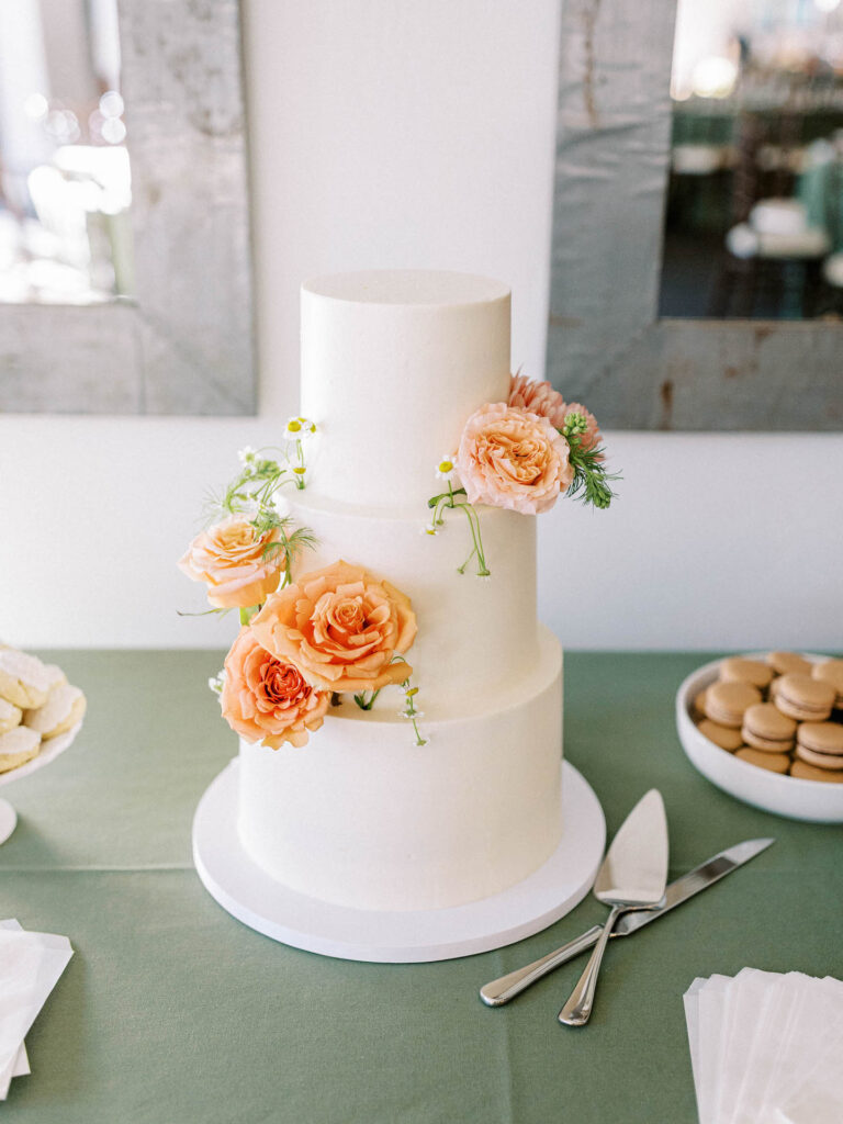 Three tired white wedding cake with peach colored flowers and chamomile on desert table.