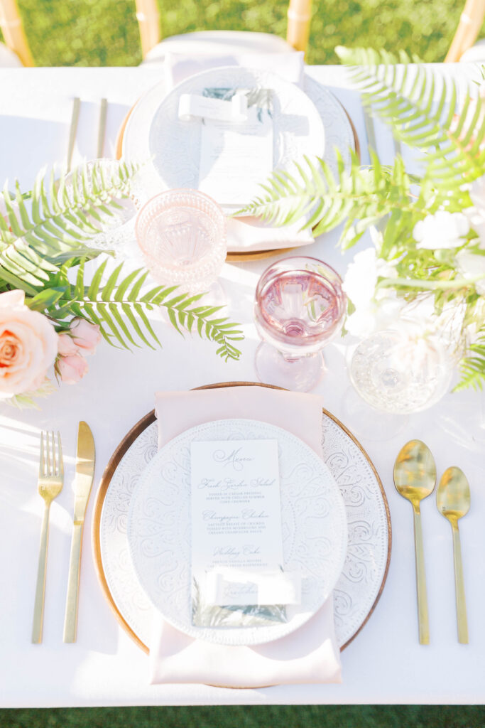 Tropical wedding reception table place setting with ferns and pink glasses.
