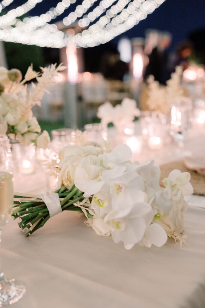 A bouquet of white flowers on a white table and more white tables and lights in the background.
