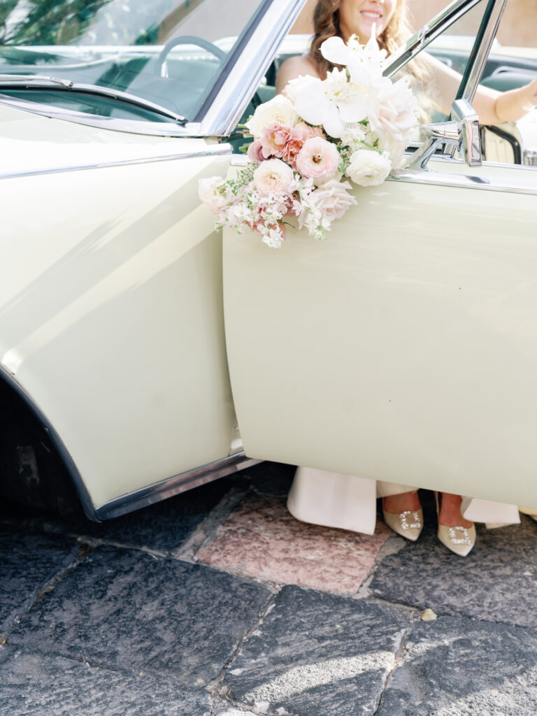 Bride sitting in driver's seat of vintage car holding bridal bouquet.