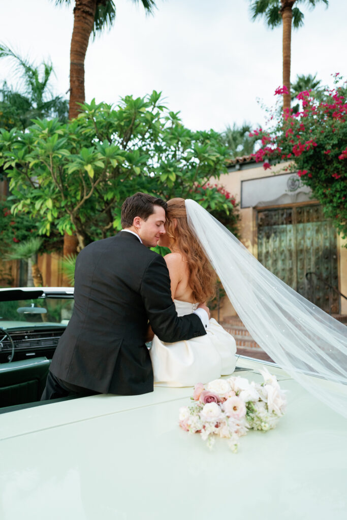 Groom with arm around bride sitting on top of back seat of vintage car with bouquet behind them on trunk.
