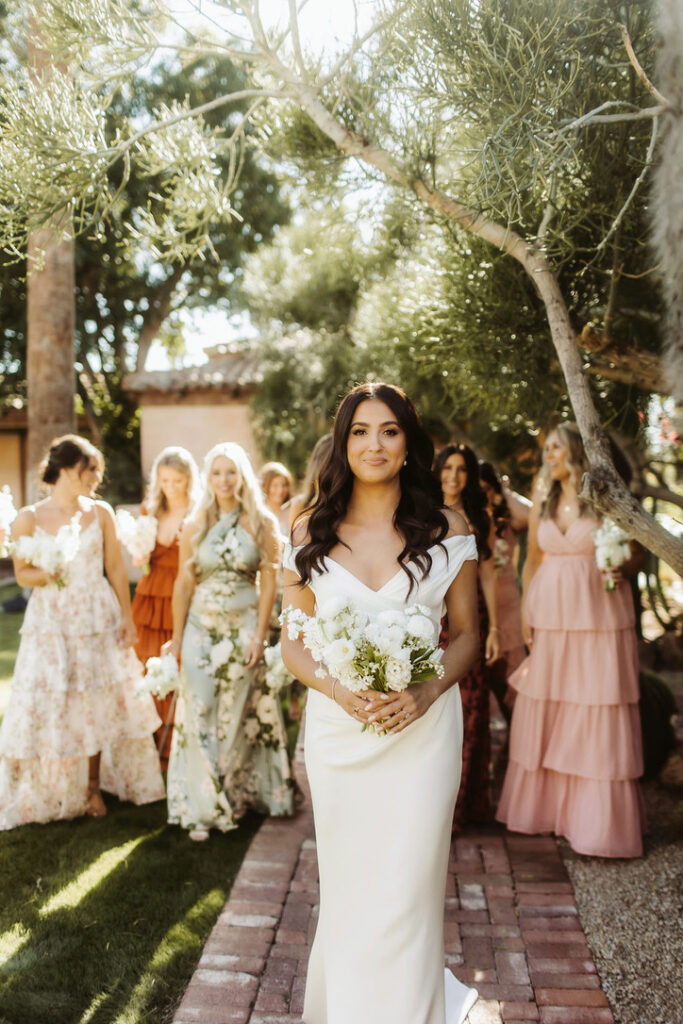 Bride standing in front of bridesmaids in different pattern and colored dresses of light pink, green, and copper.