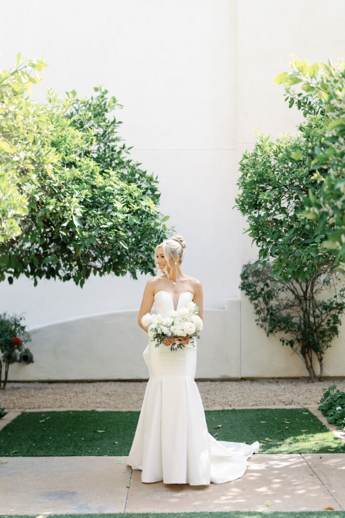 Bride standing in front of white wall and trees, looking to side while holding bouquet of white flowers.