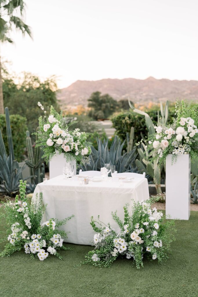Wedding reception sweetheart table in desert landscape space with two ground floral arrangements in front of table, and two floral arrangements on pillars behind the table.