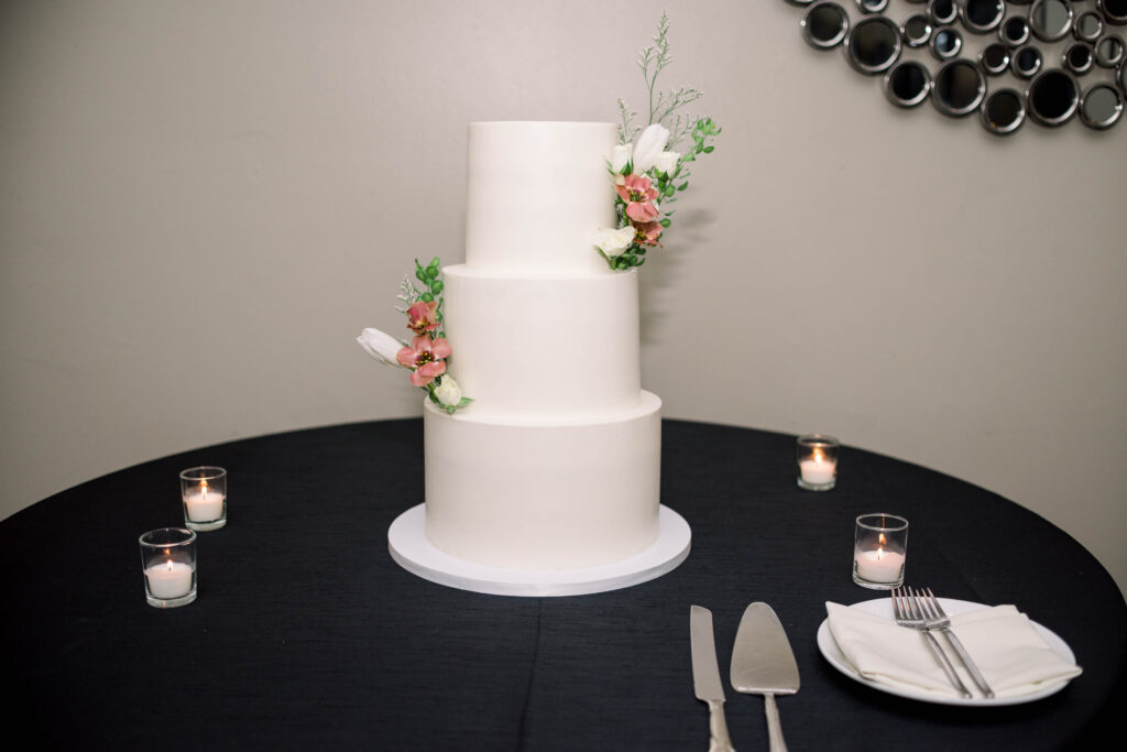 Three tiered white wedding cake with white and pink flowers and greenery added to top two tiers sitting on black linens round table.