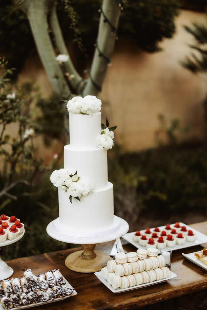Three tiered white wedding cake with flowers added and assorted deserts on table below.