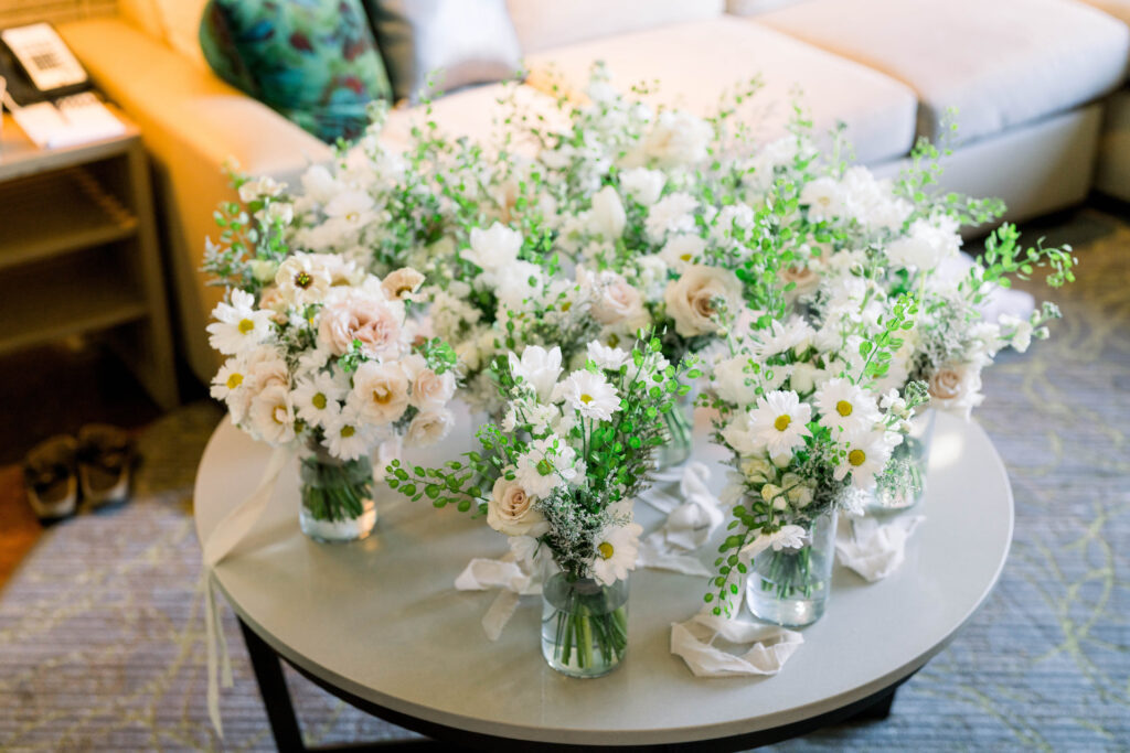 Round table filled with glass vases of bridesmaid bouquets of white flowers, some with yellow centers, and blush flowers.