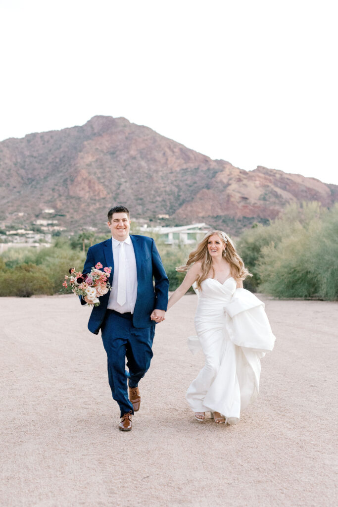 Bride and groom walking while holding hands in desert landscape area, groom holding bride's bouquet.