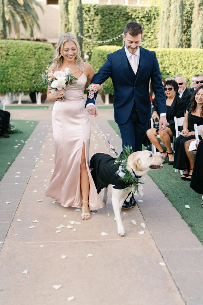 Bridesmaid and groomsmen walking down the aisle during ceremony holding a dog with a tux and greenery collar on by leash.