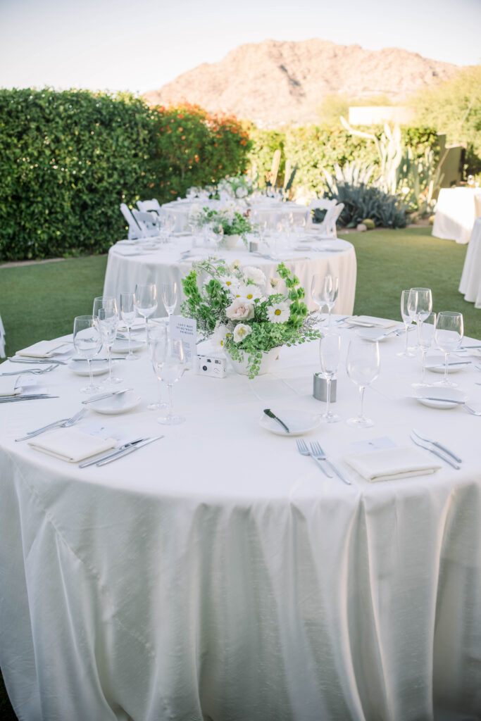 Outdoor reception space with round tables with white linens and white floral and greens centerpiece.