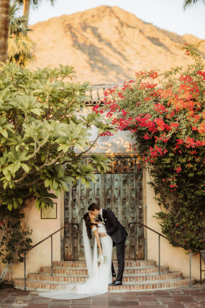 Bride and groom kissing on steps of Royal Palms large double doors outside and mountain view in distance.