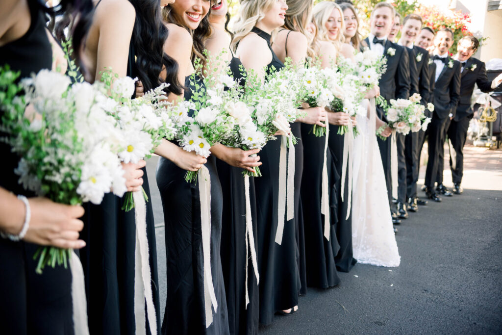 Wedding party in a line with bride and groom in center, bridesmaids and groomsmen in black with women holding bouquets.