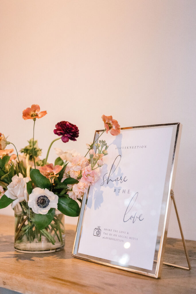 Floral arrangement in glass vase next to sign at wedding on wood table.