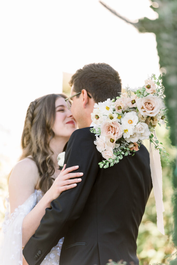 Bride and groom touching noses while embracing with bride holding her bouquet over groom's shoulder behind his neck.