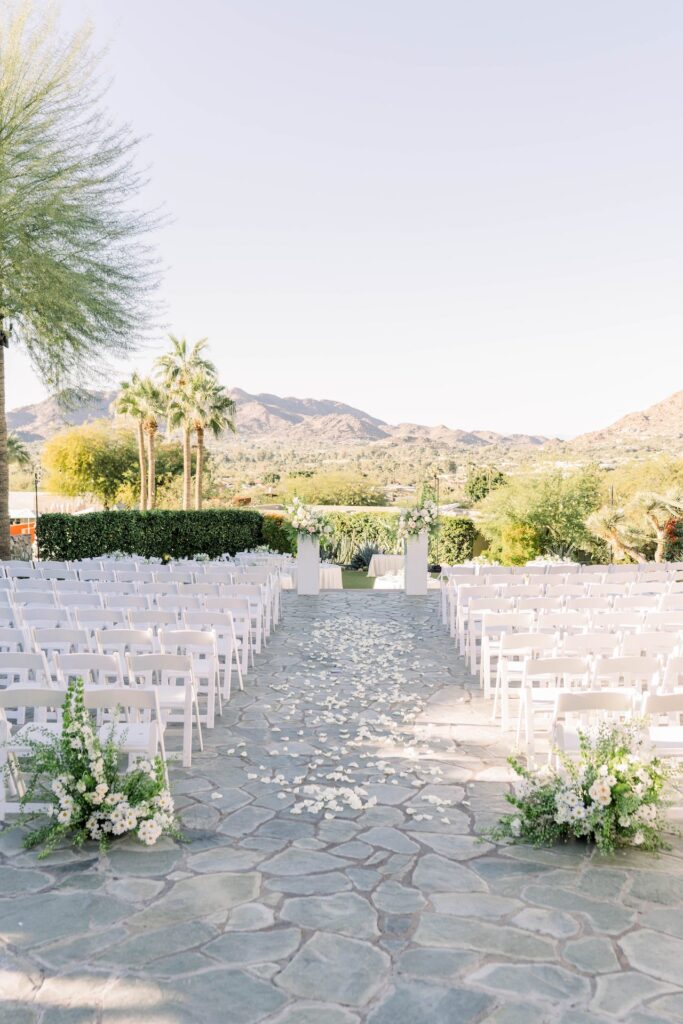 Outdoor wedding reception space with large stone ground, back of aisle ground flower arrangements, rose pedals down the aisle and flowers on low pillars in the altar space.