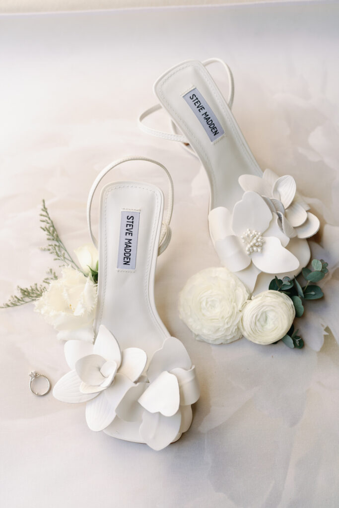 Bride's shoes of white flowers next to ring and real white flowers with greenery.