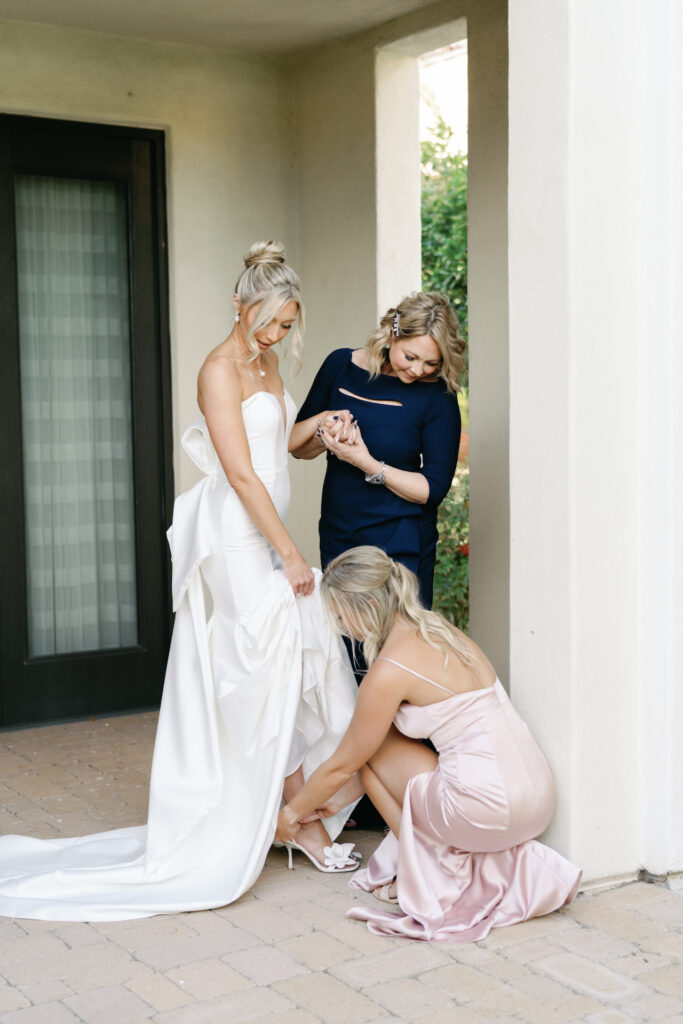 Bridesmaid helping bride to get shoe on with another woman standing next to bride.