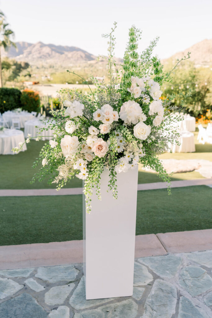 Large floral arrangement of white flowers and greenery on a white mid-height column.