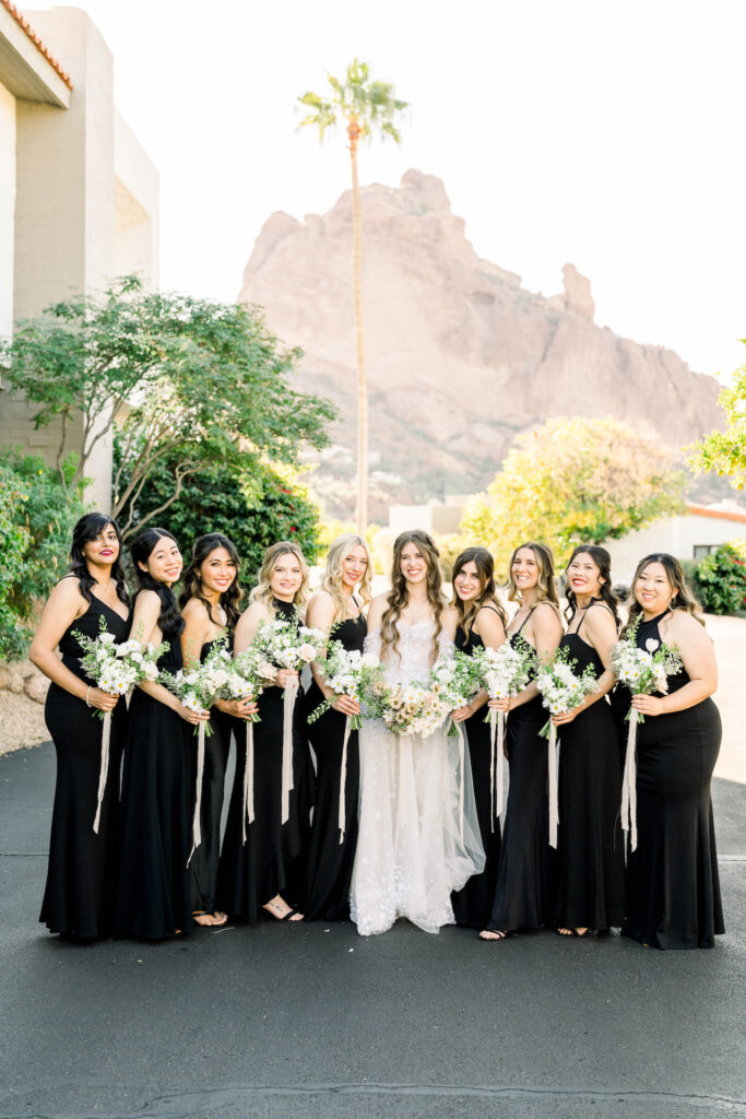 Bride standing with bridesmaids in a line wearing black dresses and holding bouquets of white flowers with greenery with Camelback mountain distance.