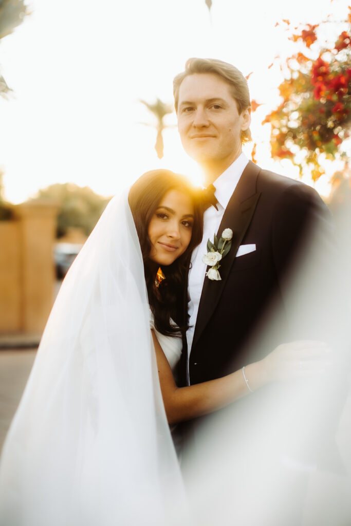 Sun setting behind bride and groom, with bride's head resting on groom's chest and both smiling.
